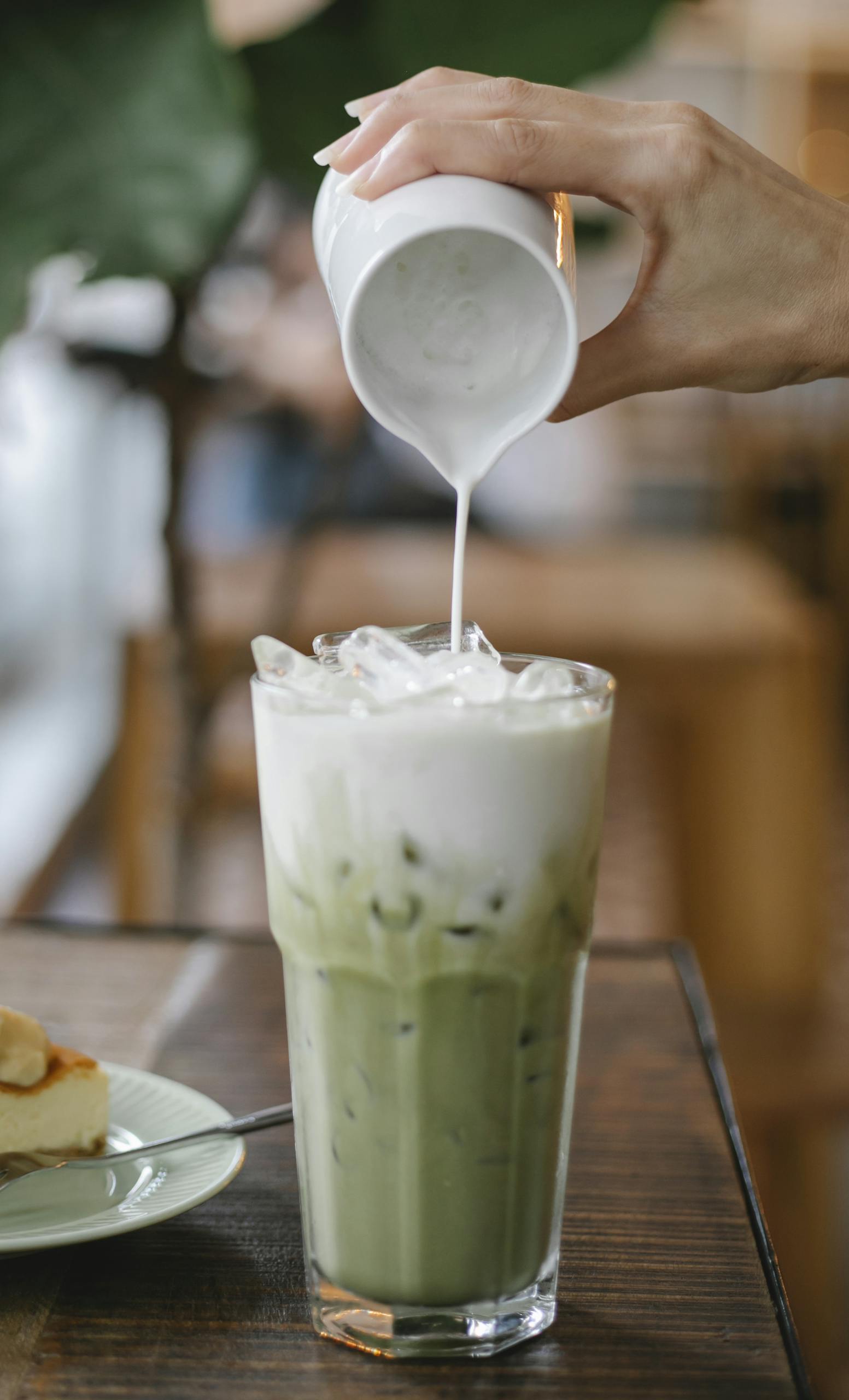 Crop unrecognizable female barista adding fresh milk from jigger into glass of yummy iced matcha latte